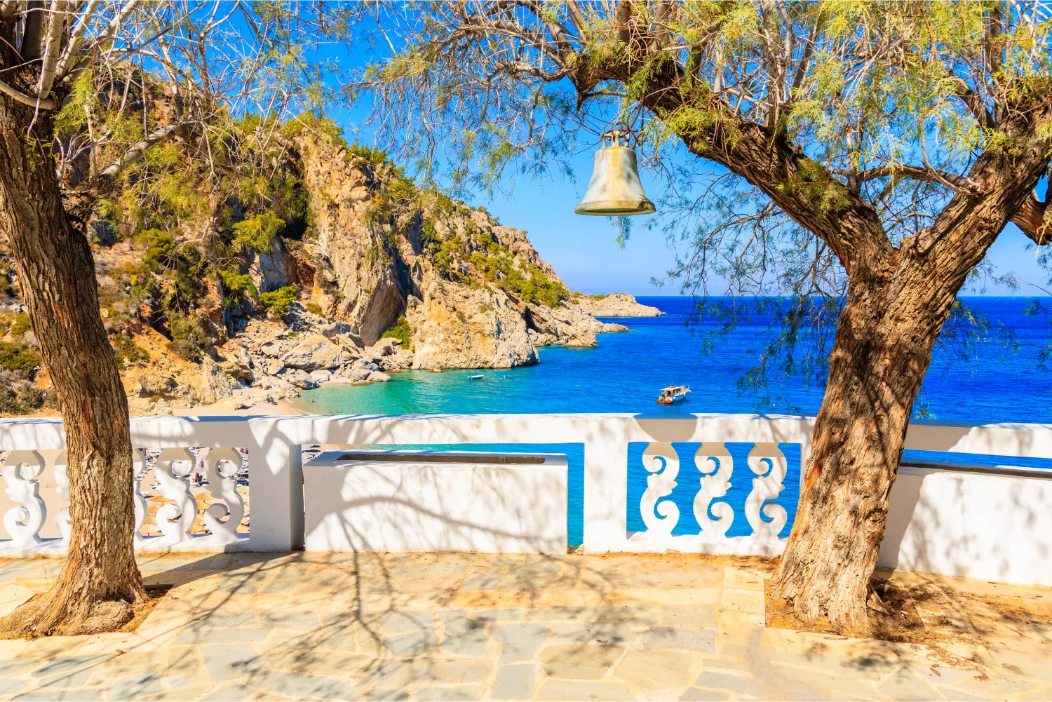 Cloche accrochée à un arbre et vue sur la plage de Kyra Pynagia depuis la terrasse d'une église à Karpathos