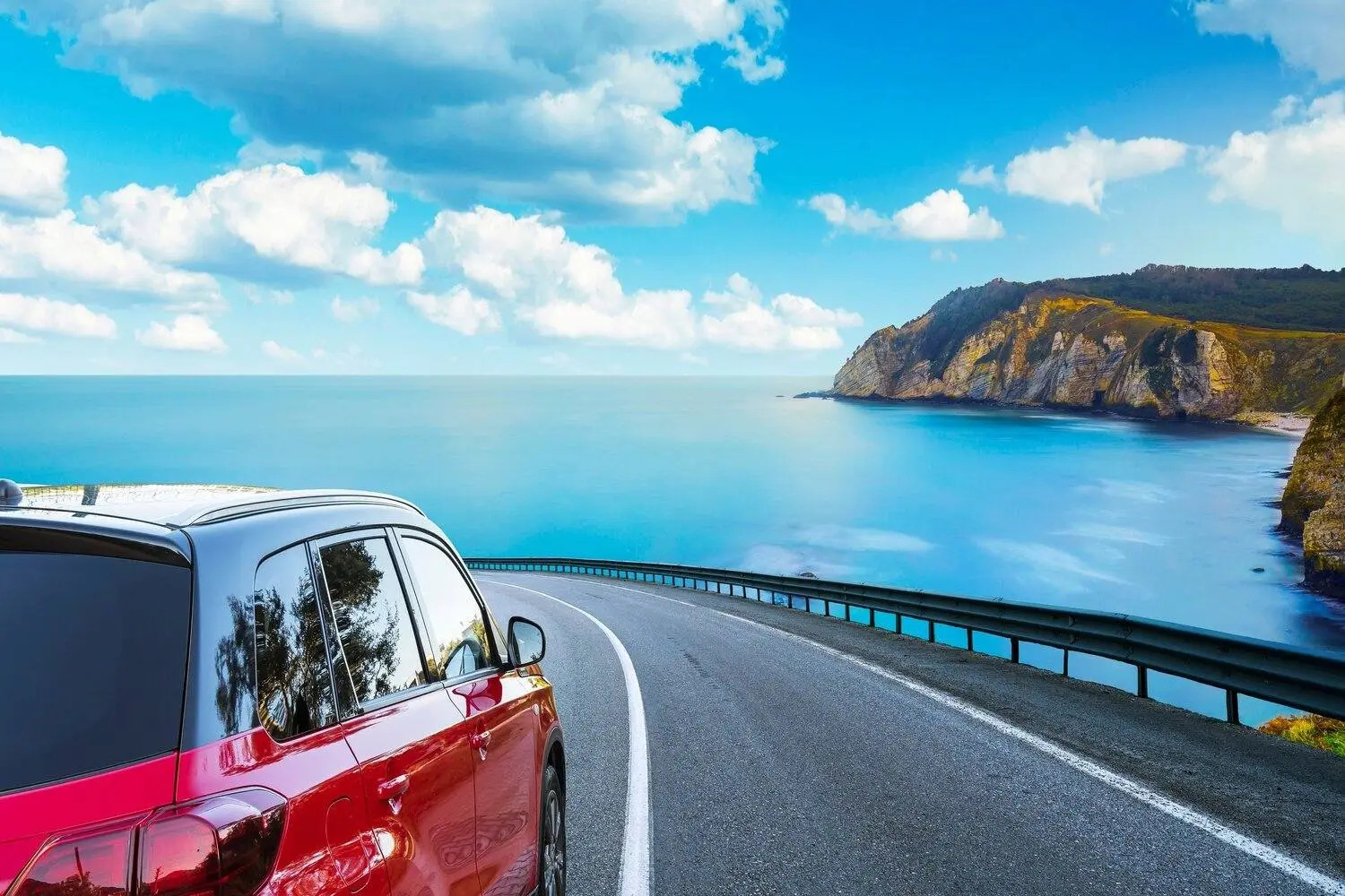 Voiture rouge roulant sur une route côtière surplombant l'océan bleu et les falaises rocheuses sous un ciel lumineux avec des nuages