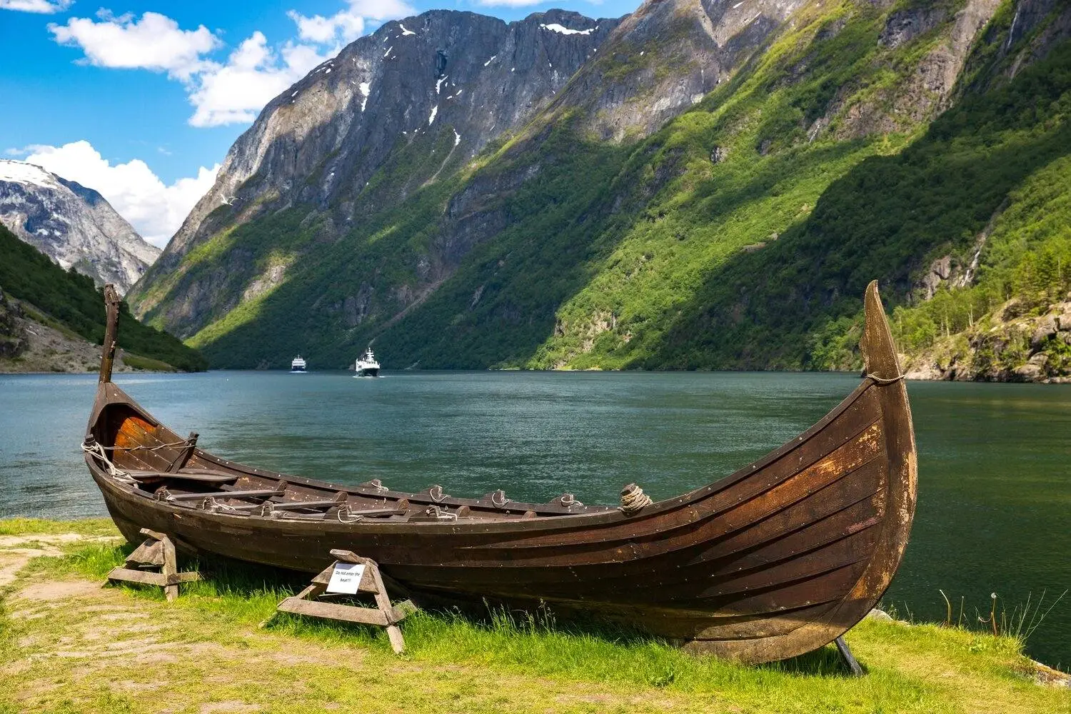 Un navire viking en bois repose sur la rive herbeuse d'un fjord calme, entouré de montagnes verdoyantes escarpées et de falaises rocheuses.