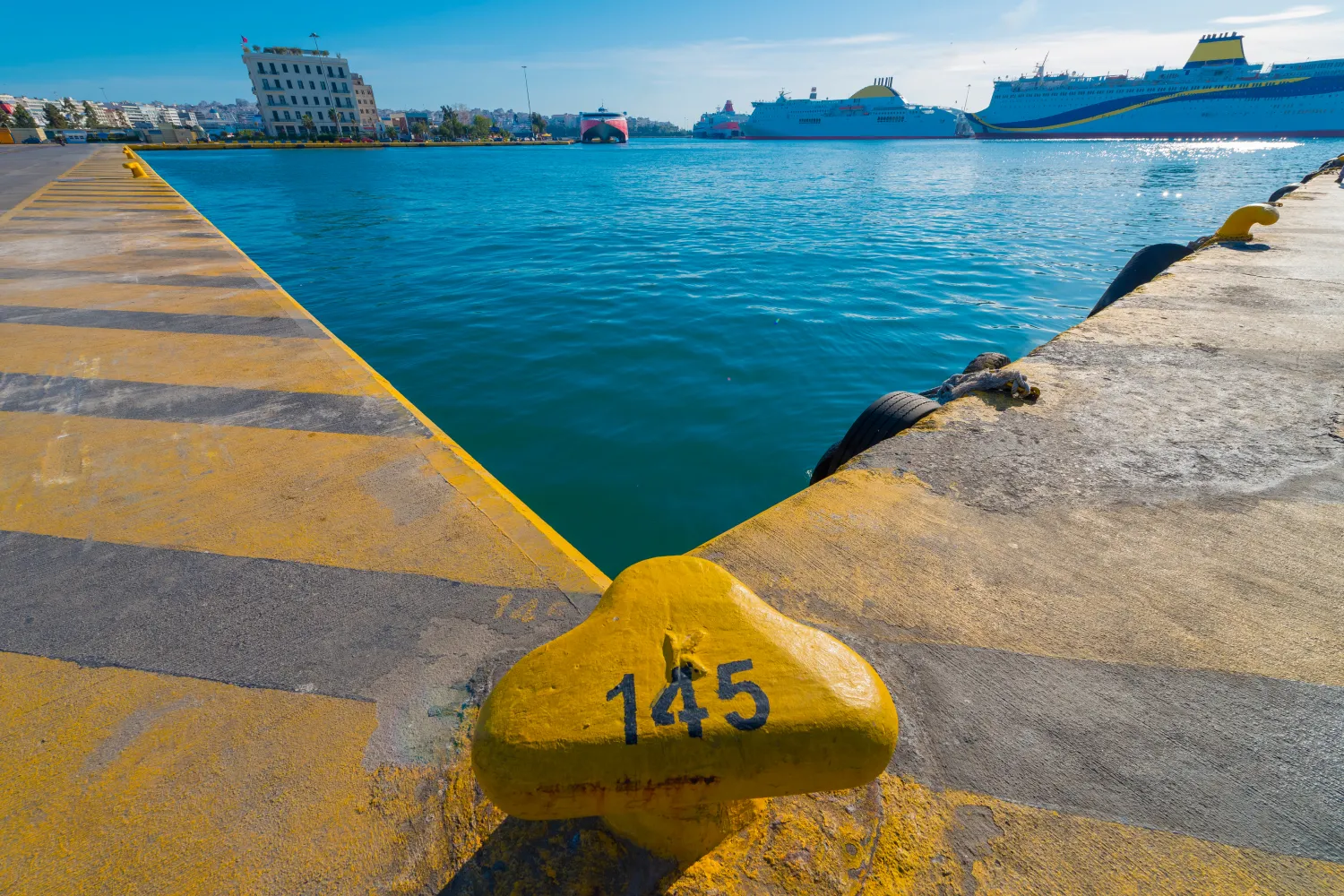 Le port du Pirée en Grèce montre de grands ferries à passagers amarrés au quai avec une borne d'amarrage jaune numérotée 145 au premier plan sous un ciel dégagé.